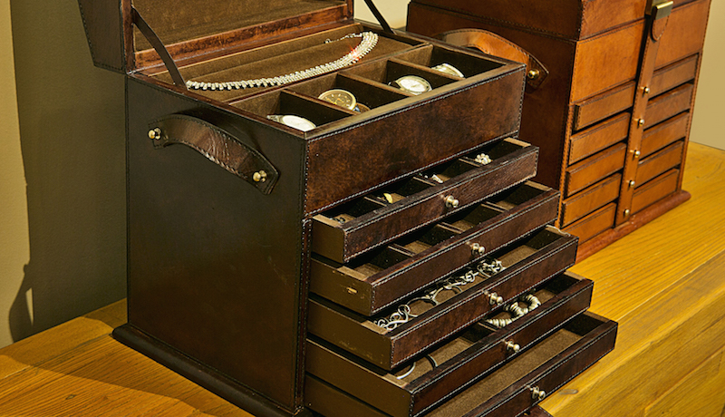 A caixa de joias mais alta tem cinco gavetas com divisões variadas, nichos para relógios e uma almofada para apoiar a joia mais usada. As alças de couro ajudam no transporte.