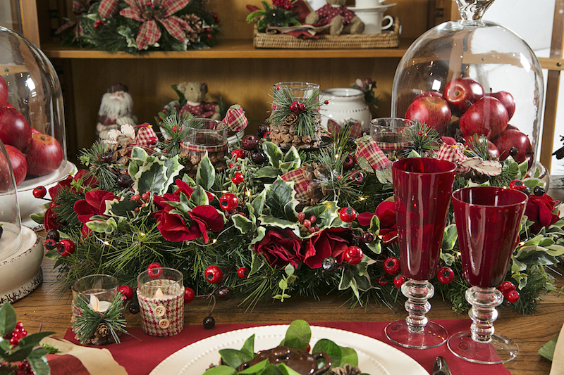 O centro de mesa tem porta-velas feiots de copinhos de vidor forrados com pinhas, e é feito de ramos natalinos com azevinho e flores vermelhas.