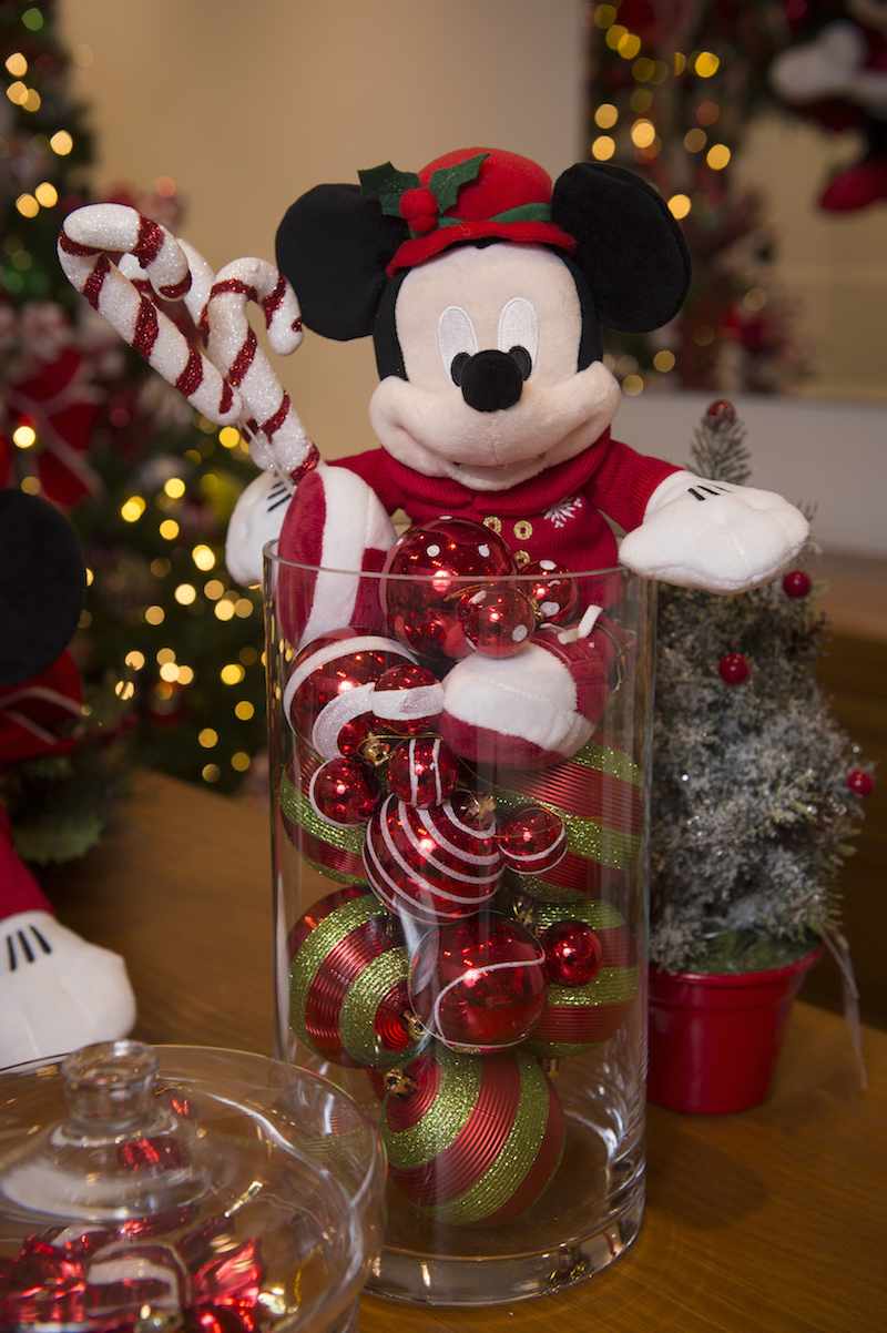 Enquanto as crianças comem, o Mickey guarda um cilindo de vidro cheio de bolas com orelhas, e segura um ramo com bengalinhas de Natal.