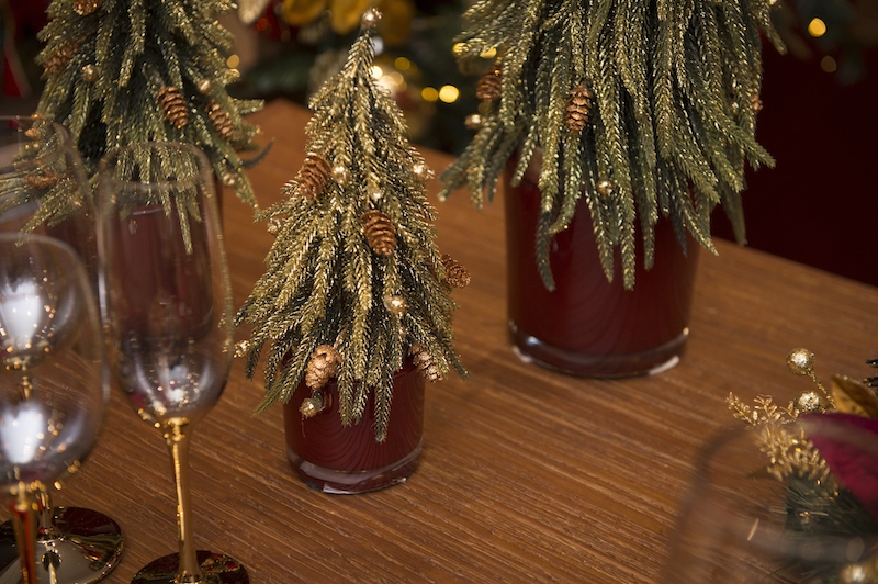 Mini árvores cobertas de "neve dourada" encaixam perfeitamente nos cachepots de vidro vermellho, preenchendo o centro da mesa.