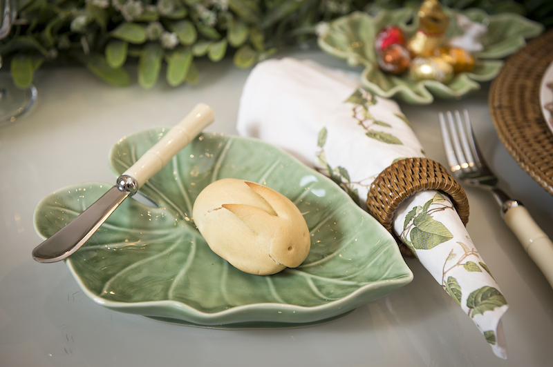 Ao lado de cada prato, uma folha de cerâmica faz o papel de pratinho de pão. O guardanapo de pano é forrado com um de papel combinando com a louça, aumentando a sensação de jardim da mesa.